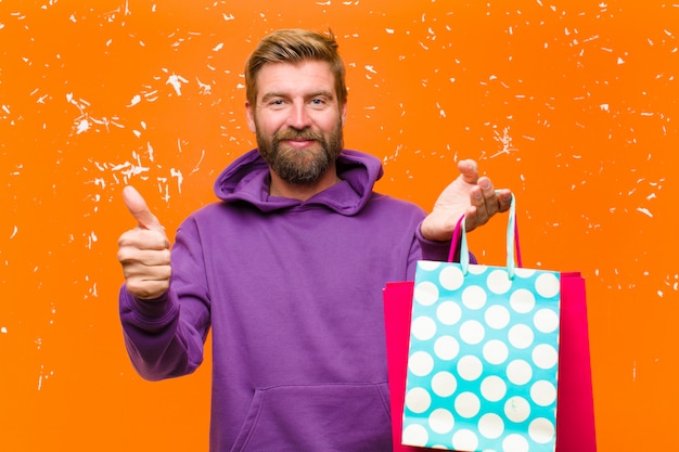 Foto giovane uomo biondo con i sacchetti della spesa che indossa una maglia con cappuccio porpora contro la parete arancio danneggiata
