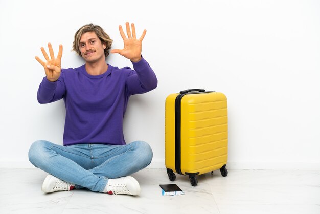 指で9を数える床に座っているスーツケースを持つ若いブロンドの男