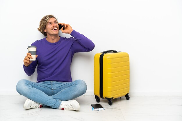 Young blonde man with suitcase sitting on the floor holding coffee to take away and a mobile