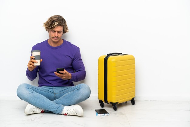 Young blonde man with suitcase sitting on the floor holding coffee