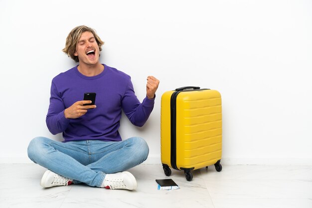 Young blonde man with suitcase sitting on the floor with phone in victory position