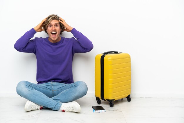 驚きの表情で床に座っているスーツケースを持つ若いブロンドの男