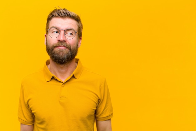 Young blonde man wondering, thinking happy thoughts and ideas, daydreaming, looking to copy space on side against orange wall