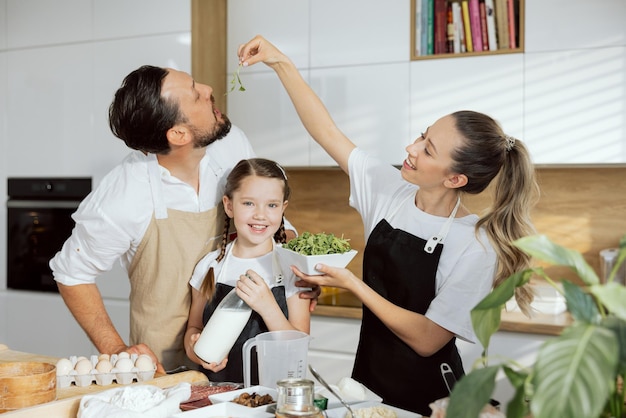 Young blonde mother feeds dad with arugula from her hands Delighted curious daughter with braids