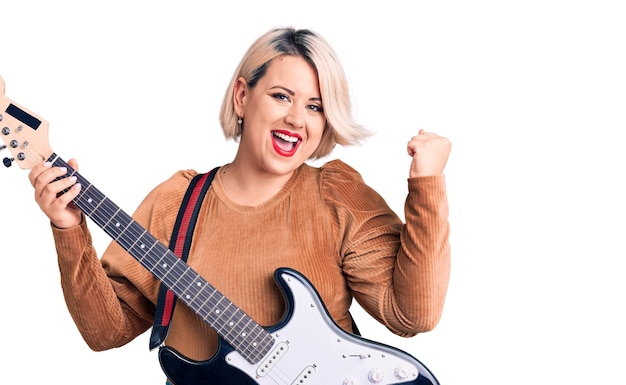 Young blonde plus size woman playing electric guitar screaming proud, celebrating victory and success very excited with raised arms