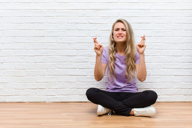 Young blonde pretty woman crossing fingers anxiously and hoping for good luck with a worried look