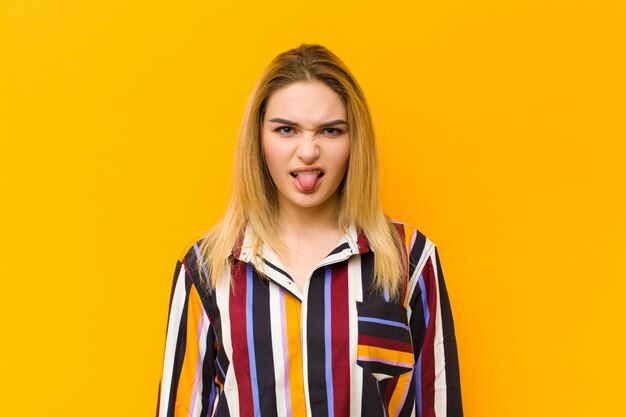 Young blonde pretty woman feeling disgusted and irritated, sticking tongue out, disliking something nasty and yucky against orange wall