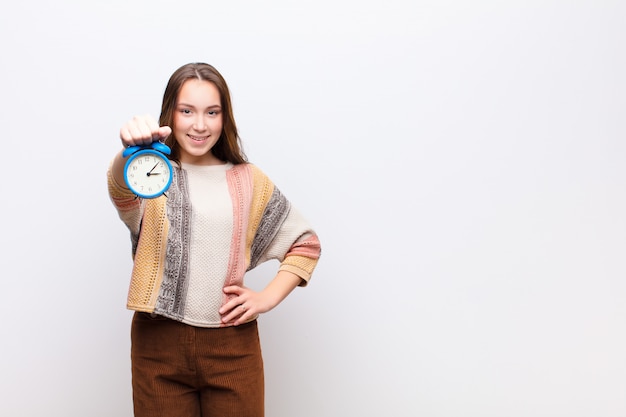 Young blonde pretty woman holding an alarm clock against white wall