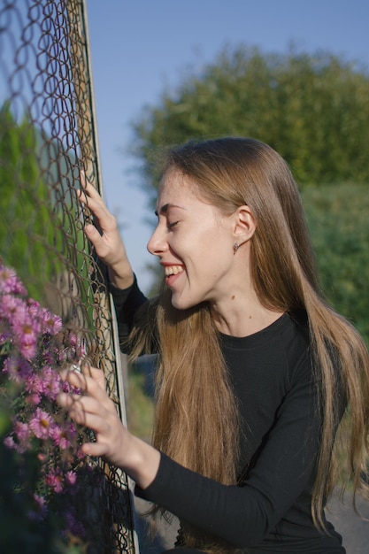 Young blonde romantic girl looks at purple flowers through the fence and touches the flowers.