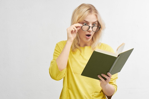 Young blonde in round glasses reads a book with a surprised face in yellow clothes. Isolated in white studio.