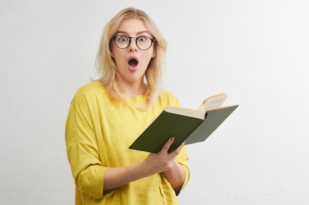 Young blonde in round glasses reads a book with a surprised face in yellow clothes. Isolated in white studio.