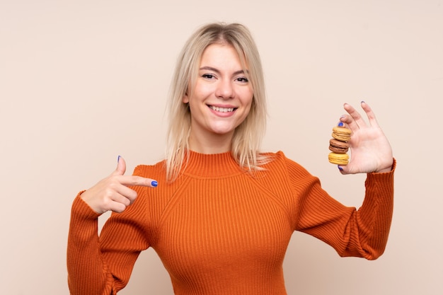 Young blonde Russian woman over isolated wall holding colorful French macarons and pointing it