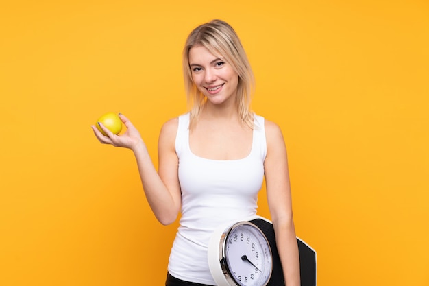 Young blonde russian woman over isolated yellow wall with weighing machine and with an apple