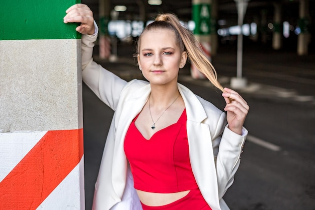 Young blonde schoolgirl in a red suit and a white jacket posing in the parking lot