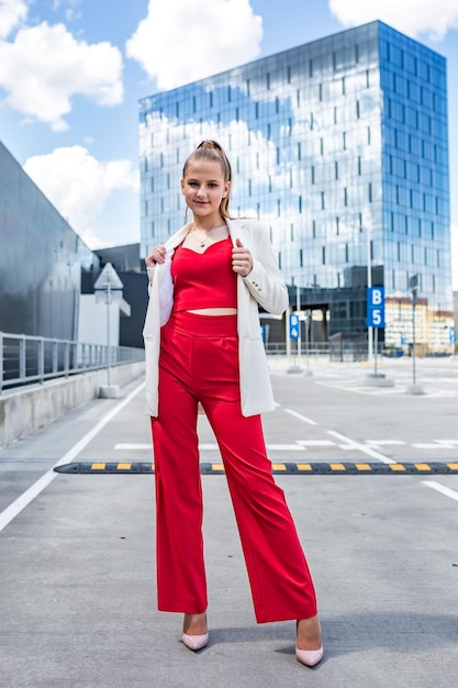 Young blonde schoolgirl in a red suit and a white jacket posing in the parking lot