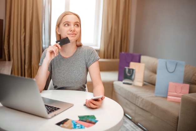   Young blonde shopper with laptop and credit card is thinking about goods while sitting at the table