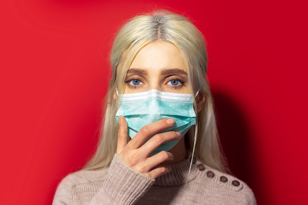 빨간 벽에 고립 된 의료 독감 마스크를 쓰고 입에 손을 잡고 파란 눈을 가진 젊은 금발 아픈 소녀.