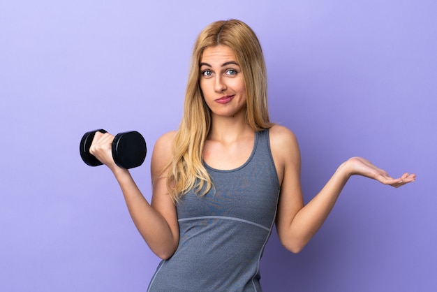 Фото Молодая белокурая женщина спорта делая поднятие тяжестей над фиолетовой стеной делая жест сомнений пока поднимающ плечи