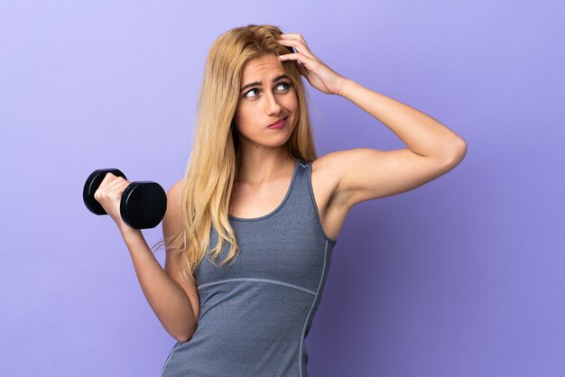 Young blonde sport woman making weightlifting over purple wall having doubts and with confused face expression