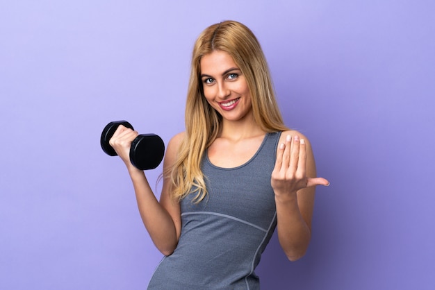 Young blonde sport woman making weightlifting over purple wall inviting to come with hand. Happy that you came