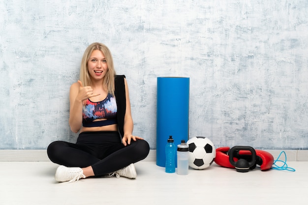 Young blonde sport woman sitting on the floor making phone gesture