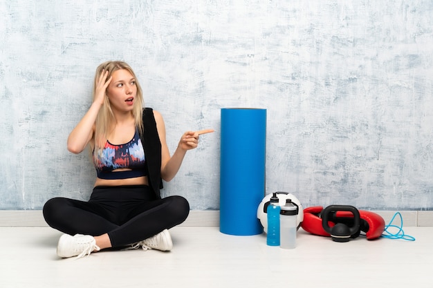 Young blonde sport woman sitting on the floor surprised and pointing finger to the side