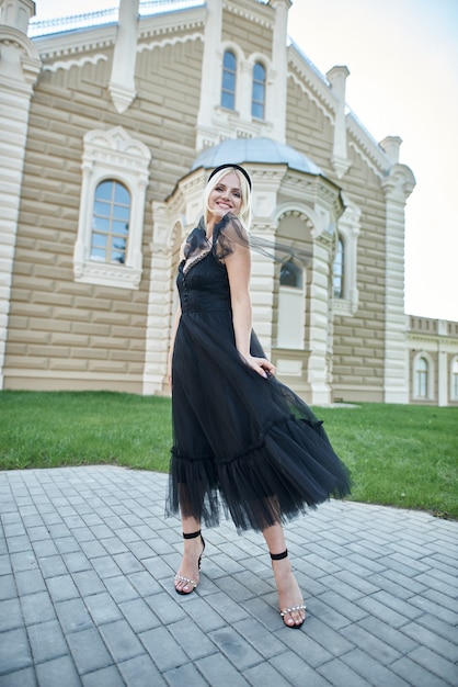 a young blonde stands near an old building in a black dress