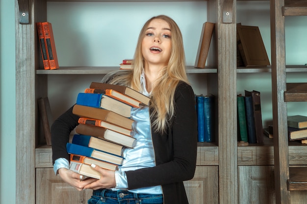 Giovane studentessa bionda con molti libri in mano e guardando la telecamera vicino a una libreria