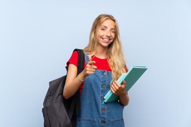 Young blonde student woman over isolated blue wall points finger at you