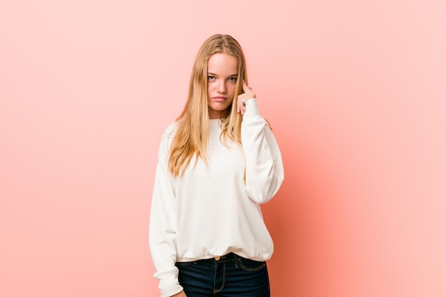 Young blonde teenager woman pointing temple with finger, thinking, focused on a task.