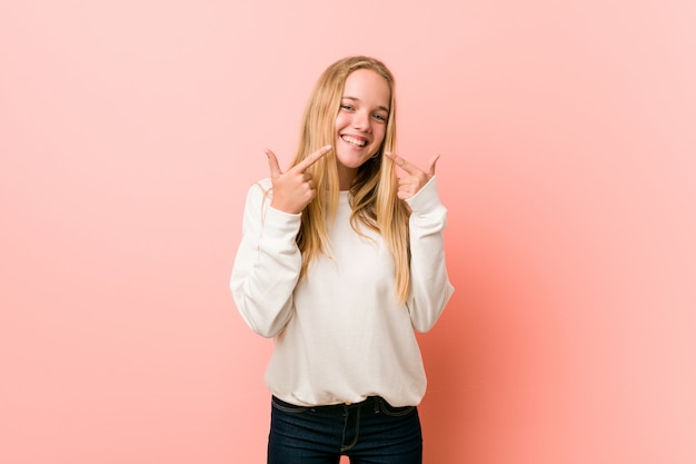 Young blonde teenager woman smiles, pointing fingers at mouth.