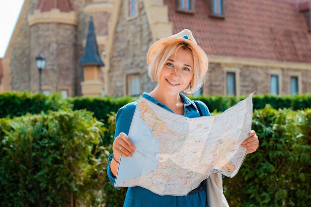 Giovane donna bionda viaggiatrice con cappello e occhiali da sole con una mappa