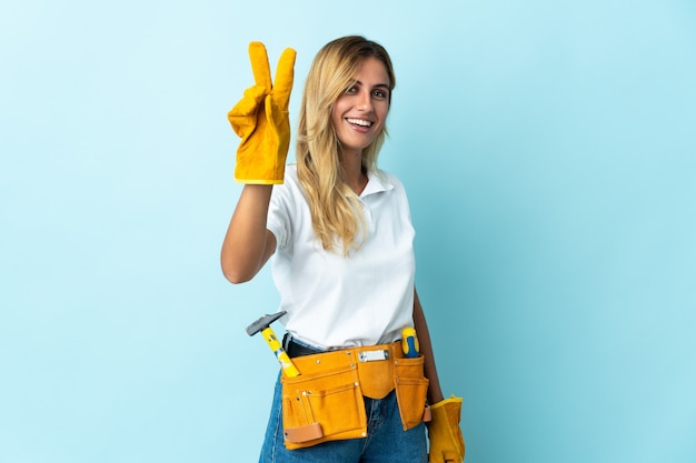 Young blonde Uruguayan electrician woman isolated on blue wall smiling and showing victory sign