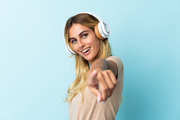 Young blonde Uruguayan isolated on blue listening music and pointing to the front