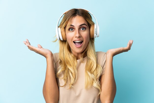 Young blonde Uruguayan isolated on blue wall surprised and listening music