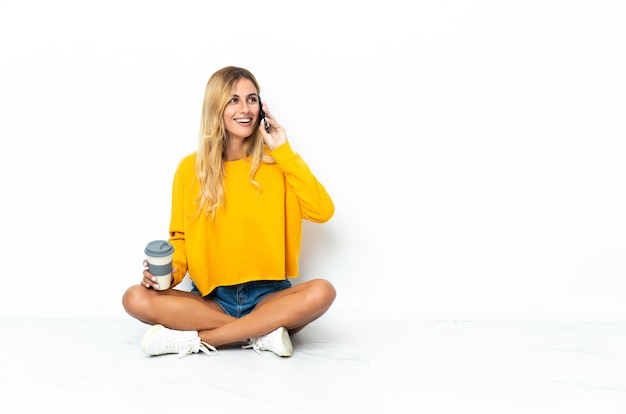 Young blonde Uruguayan woman sitting on the floor isolated on white holding coffee to take away and a mobile