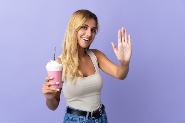 Молодая блондинка уругвайская женщина с клубничным молочным коктейлем изолирована на фиолетовом салюте рукой с счастливым выражением лица