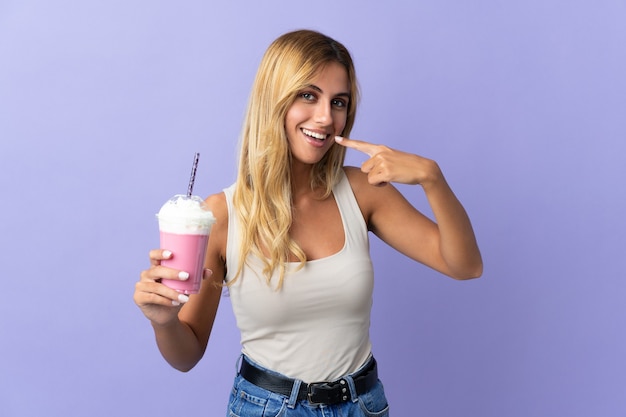 Young blonde Uruguayan woman with strawberry milkshake on purple giving a thumbs up gesture