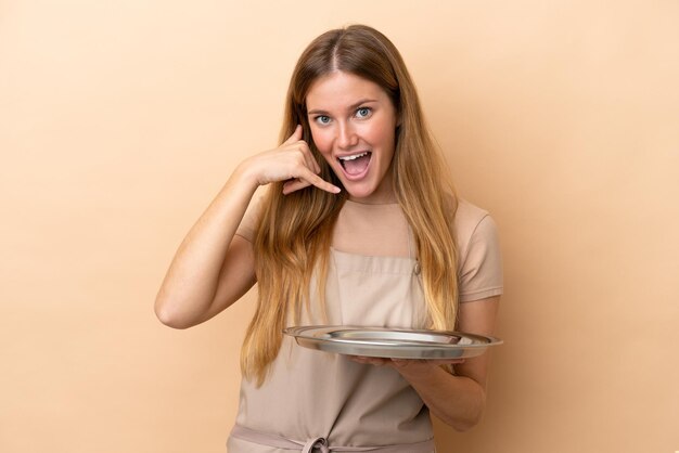 Young blonde waitress woman with tray isolated on beige background making phone gesture Call me back sign