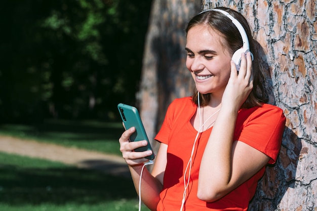 赤いシャツを着た若いブロンドは携帯電話を見て、公園で音楽を聴きます