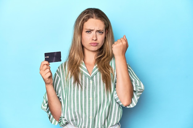 Young blonde with a credit card in blue studio showing fist to camera aggressive facial expression