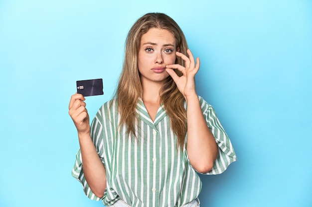 Молодая блондинка с кредитной картой в голубой студии с пальцами на губах хранит секрет