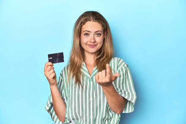 Young blonde with a credit card pointing with finger at you as if inviting come closer