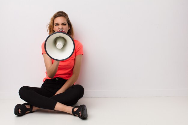 Young blonde woman  against flat color wall with a megaphone