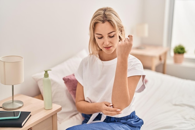 Young blonde woman applying lotion on skin at bedroom