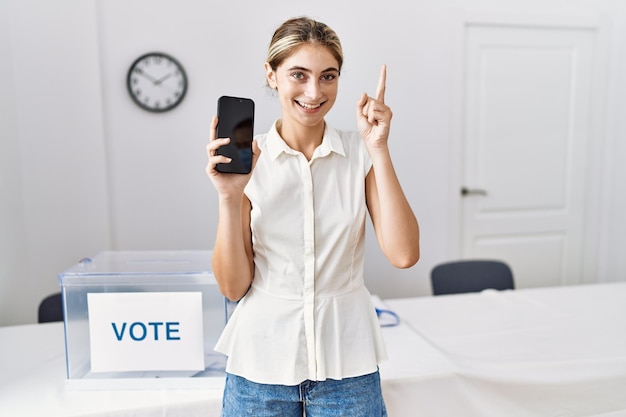 Фото Молодая блондинка на политических выборах держит смартфон, улыбаясь с идеей или вопросом, указывая пальцем со счастливым лицом номер один