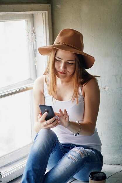 Young blonde woman in beige hat sitting on the sill and reading on her mobile phone