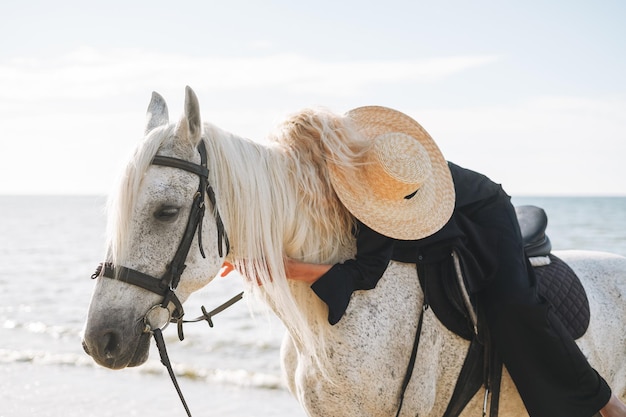 海景の背景に白い馬に乗って黒い服を着た若いブロンドの女性