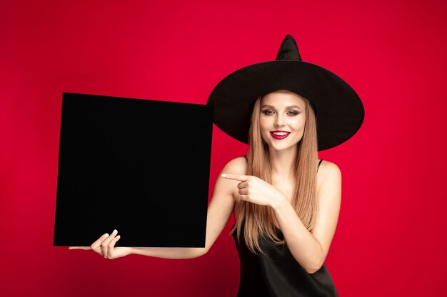 黒の帽子と赤い背景の衣装で若いブロンドの女性。魅力的な白人女性モデルのポーズ。ハロウィーン、ブラックフライデー、サイバーマンデー、販売、秋のコンセプト。コピースペース。黒いプレートを保持します。