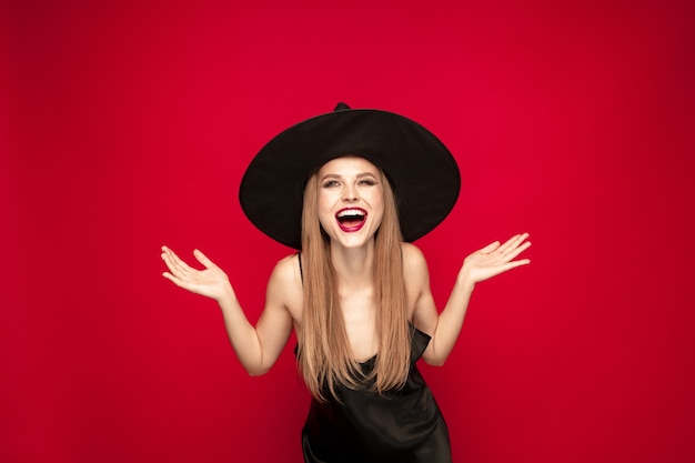 Young blonde woman in black hat and costume on red background. Attractive caucasian female model posing. Halloween, black friday, cyber monday, sales, autumn concept. Copyspace. Laughting, happy.